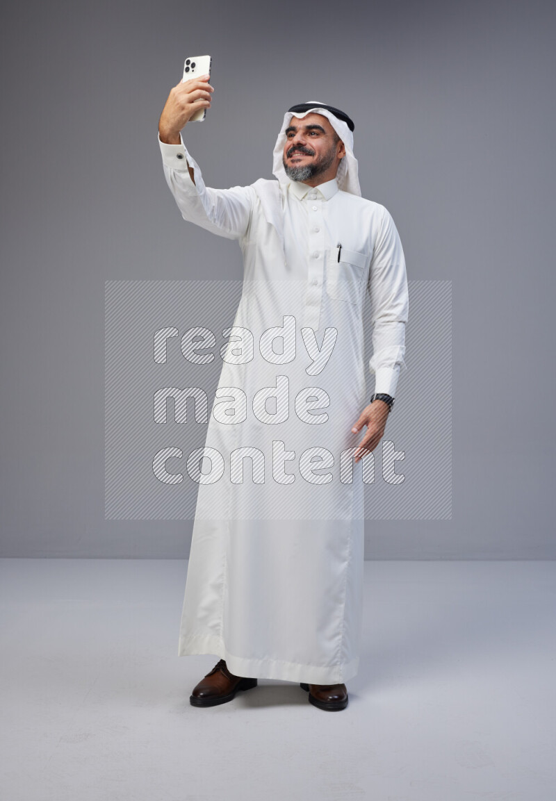 Saudi man Wearing Thob and white Shomag standing taking selfie on Gray background
