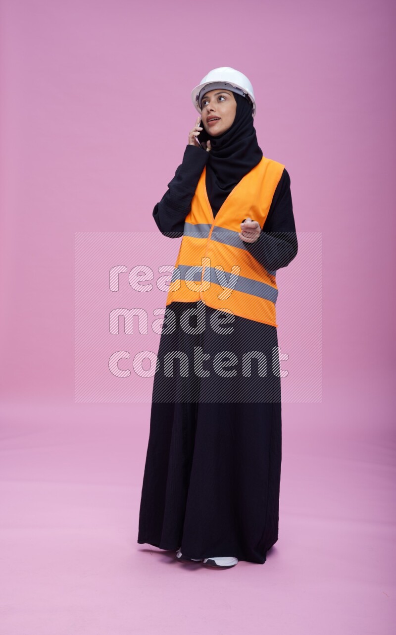 Saudi woman wearing Abaya with engineer vest and helmet standing talking on phone on pink background