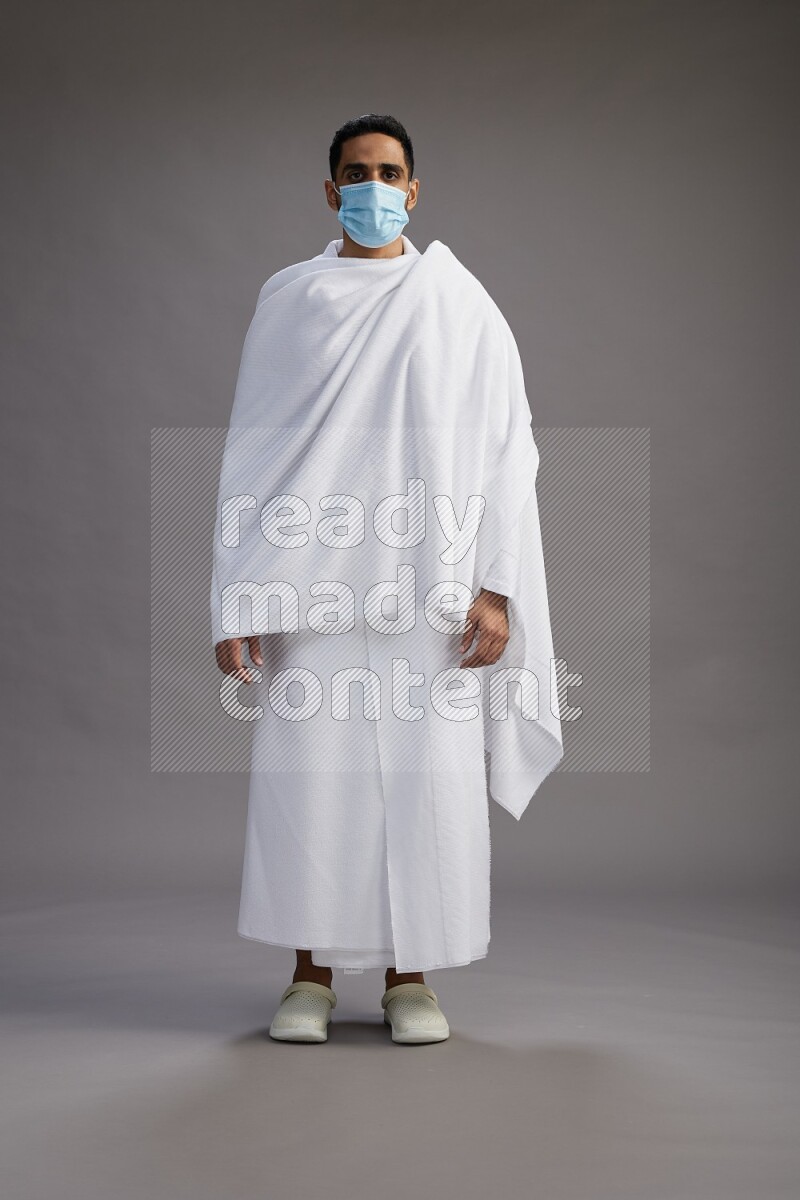 A man wearing Ehram with face mask Standing Interacting with the camera on gray background
