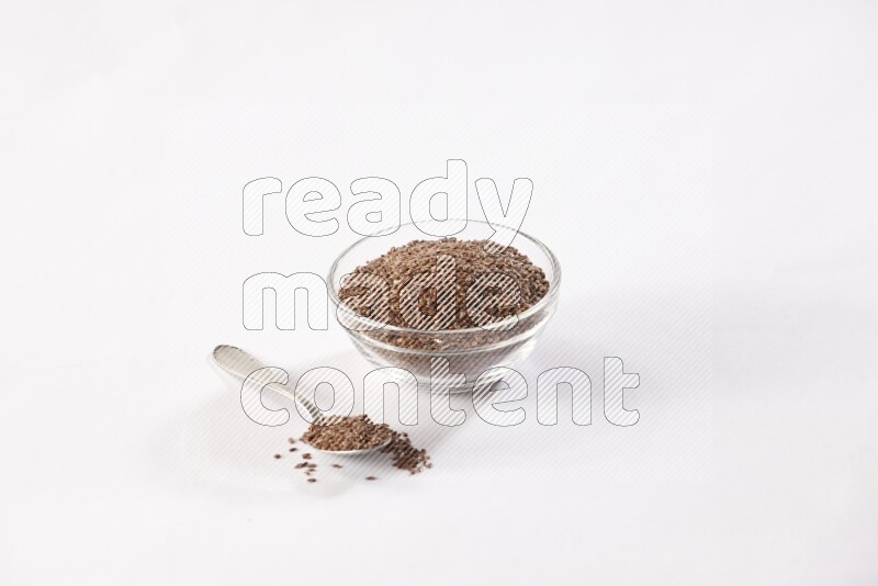 A glass bowl full of flax seeds and a metal spoon full of the seed on a white flooring