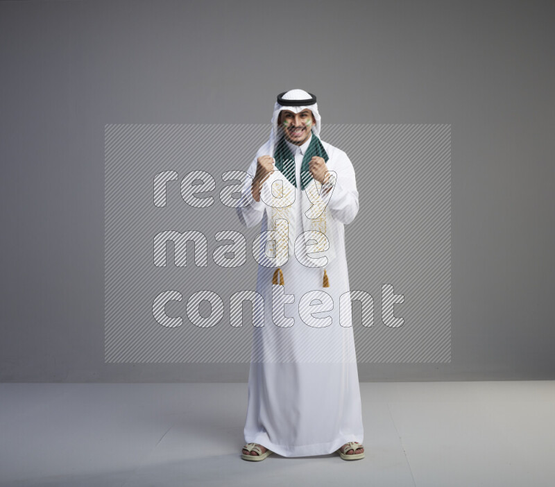 A Saudi man standing wearing thob and white shomag with face painting and Saudi flag scarf on gray background