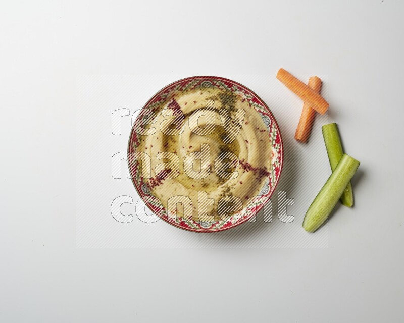 Hummus in a red plate with patterns garnished with zaatar & sumak on a white background