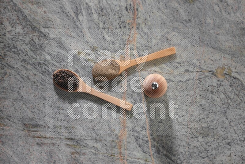 2 wooden spoons, one full of black pepper powder and the other full of black pepper beads and wooden grinder on a marble flooring