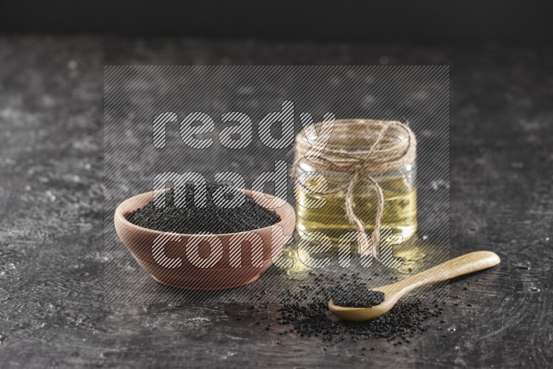 A wooden bowl and spoon full of black seeds and a glass jar of black seeds oil on a textured black flooring