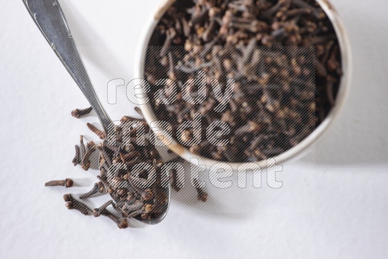 A beige ceramic bowl full of cloves and a metal spoon next to it on a white flooring