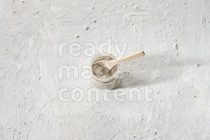 A glass jar and wooden spoon full of white pepper powder on textured white flooring