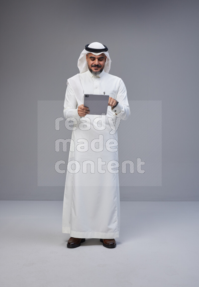 Saudi man Wearing Thob and white Shomag standing working on tablet on Gray background