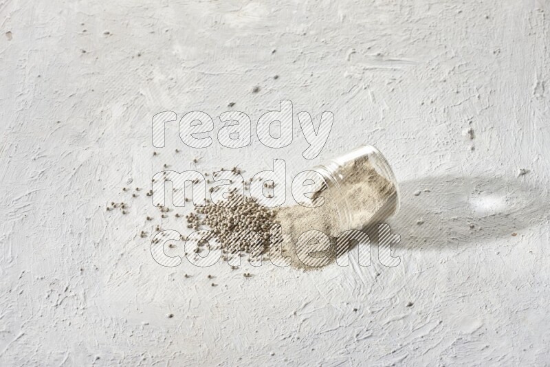 A flipped glass jar full of white pepper powder with spilled powder and white pepper beads on textured white flooring