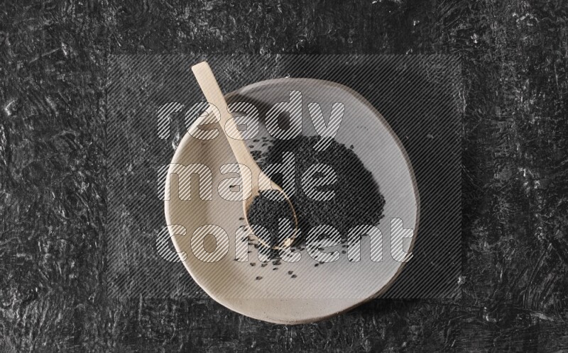 A multicolored pottery plate full of black seeds and wooden spoon full of seeds on a textured black flooring