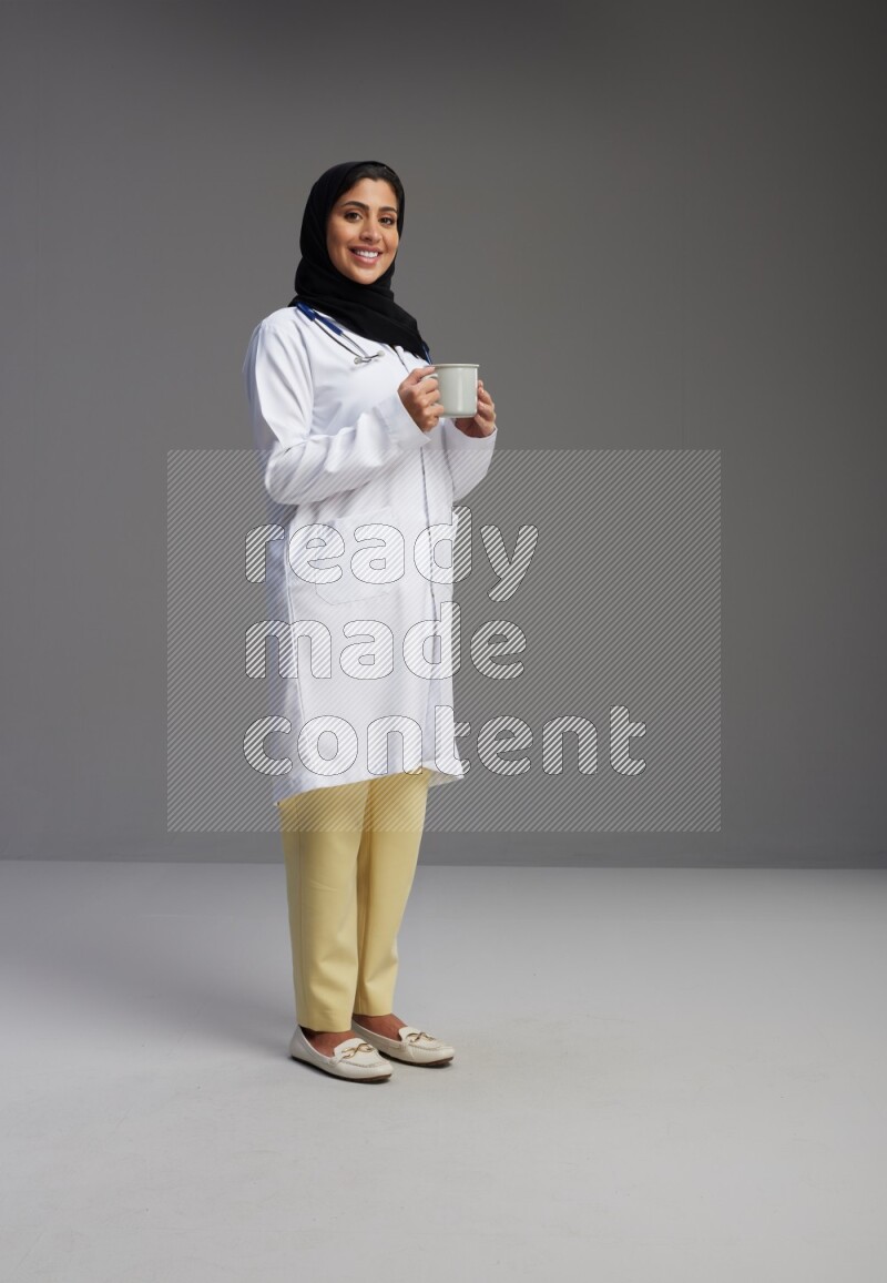 Saudi woman wearing lab coat with stethoscope standing holding mug on Gray background