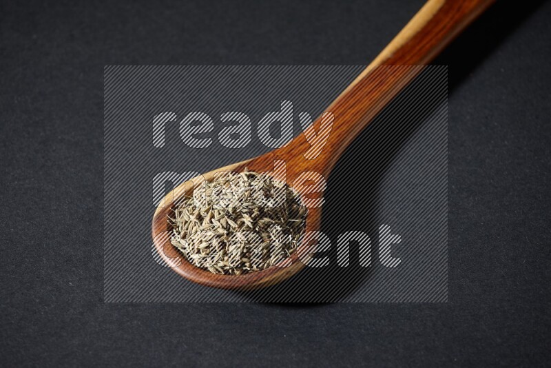 A wooden ladle full of cumin seeds on black flooring
