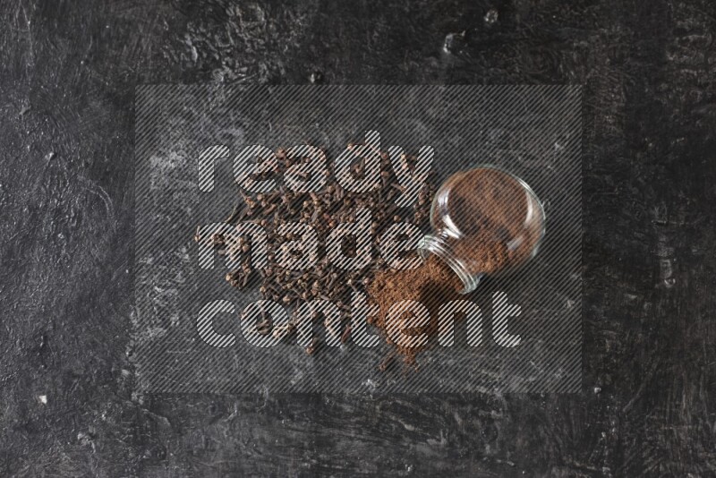 وعاء زجاجي للبهارات مقلوب ممتلئ ببودرة القرنفل والبودرة سقطت منه مع حبوب القرنفل المتناثرة على أرضية سوداء