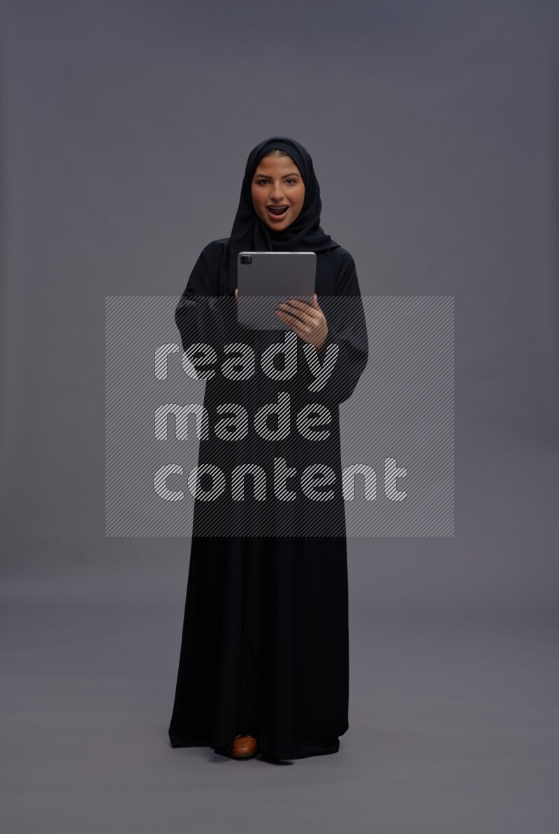 Saudi woman wearing Abaya standing working on tablet on gray background
