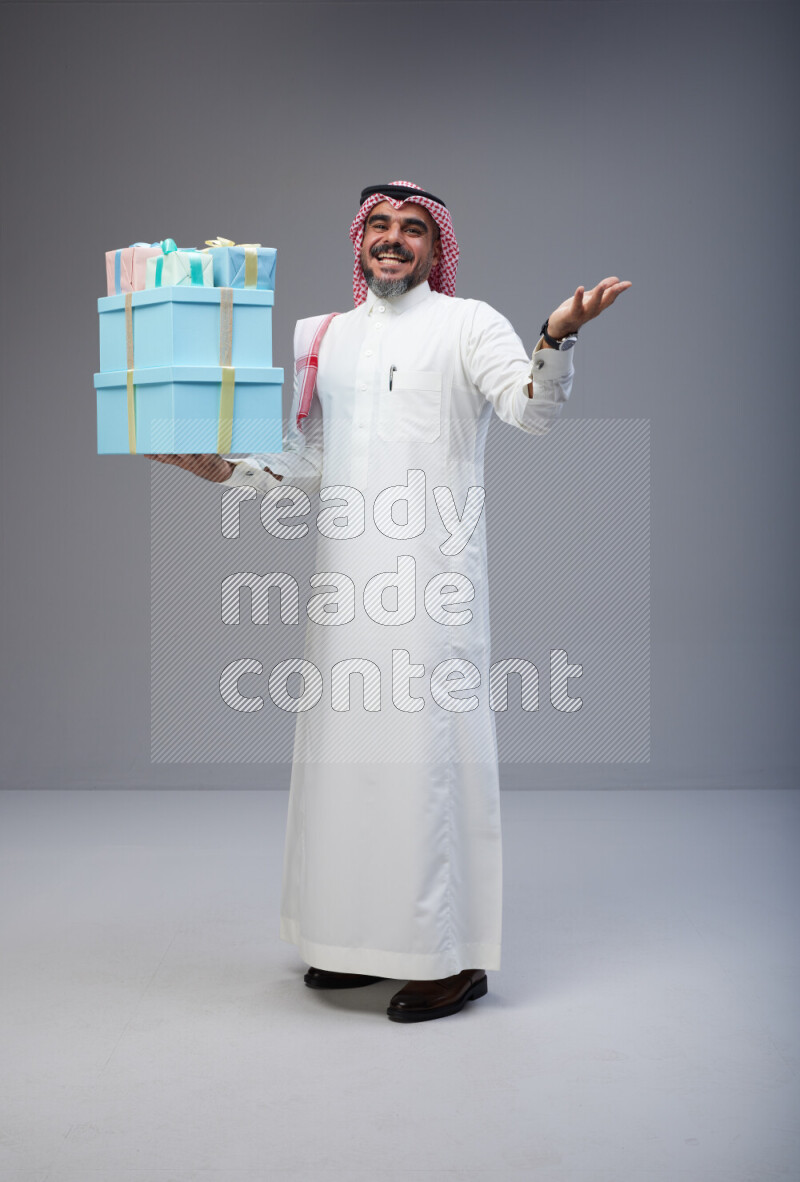 Saudi man Wearing Thob and red Shomag standing holding gift box on Gray background