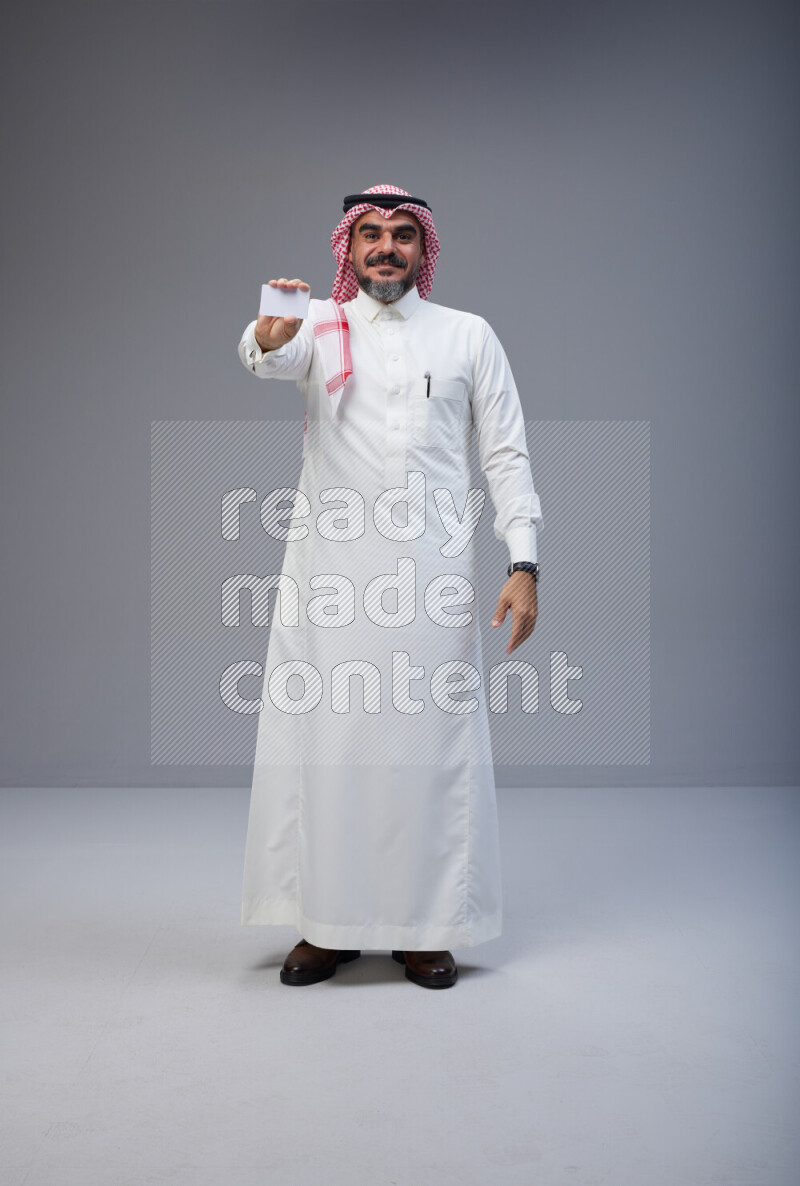 Saudi man Wearing Thob and red Shomag standing holding ATM card on Gray background