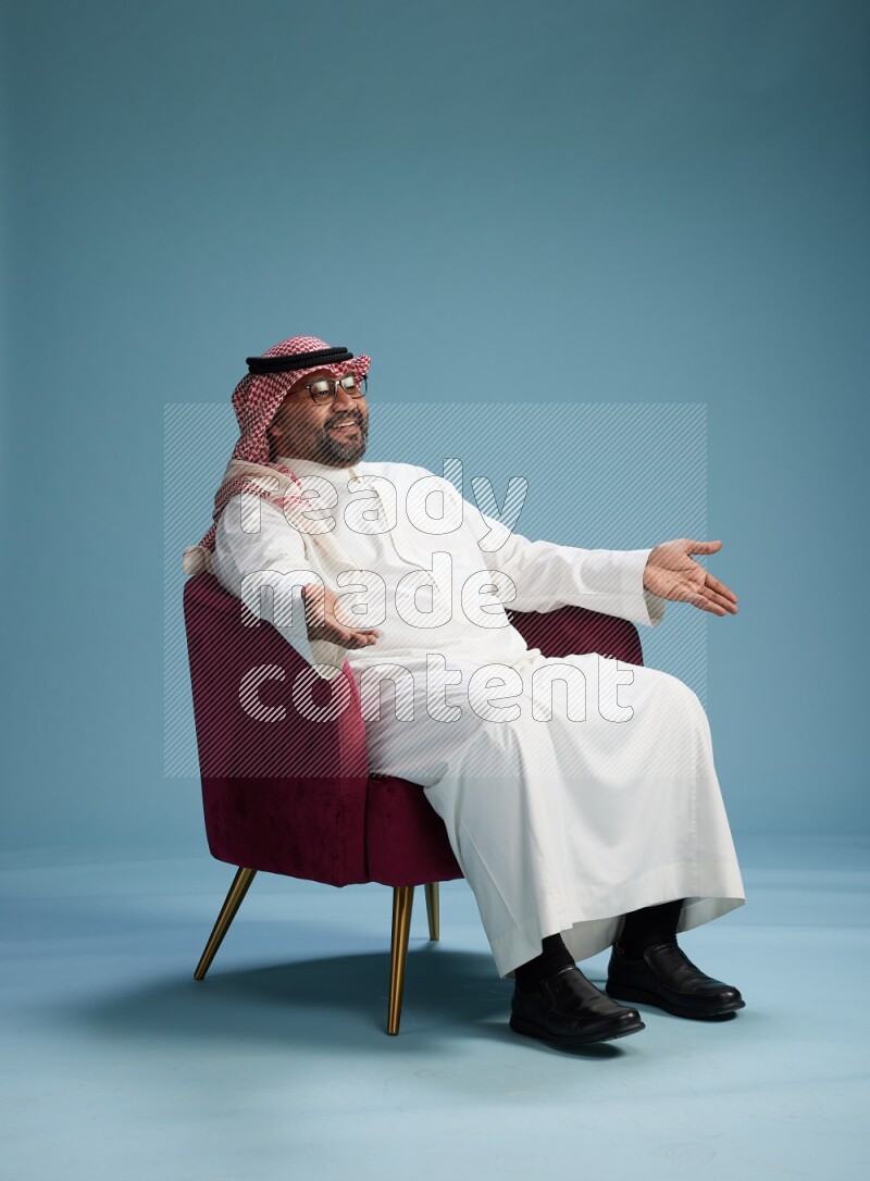 Saudi Man with shimag sitting on chair Interacting with the camera on blue background