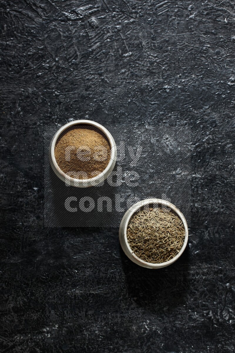 2 beige bowls, one full of cumin powder and the other full of seeds on a textured black flooring