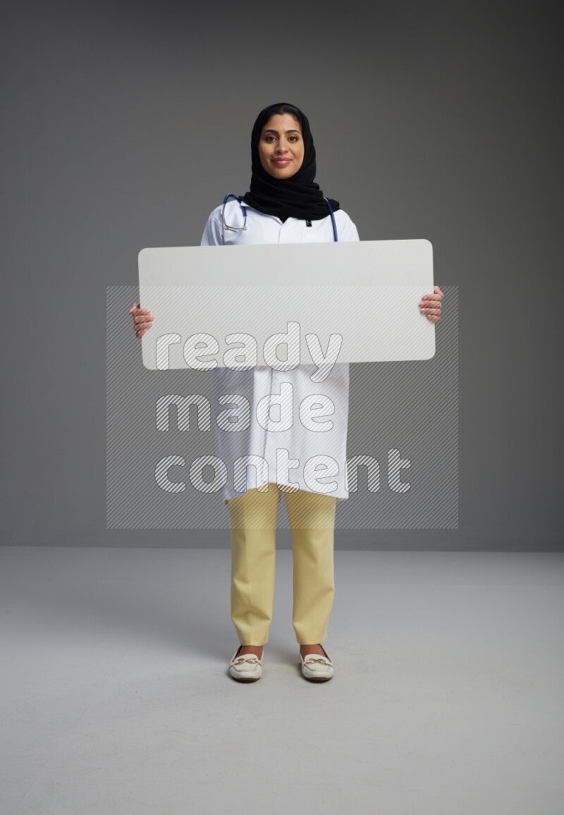 Saudi woman wearing lab coat with stethoscope standing holding board on Gray background