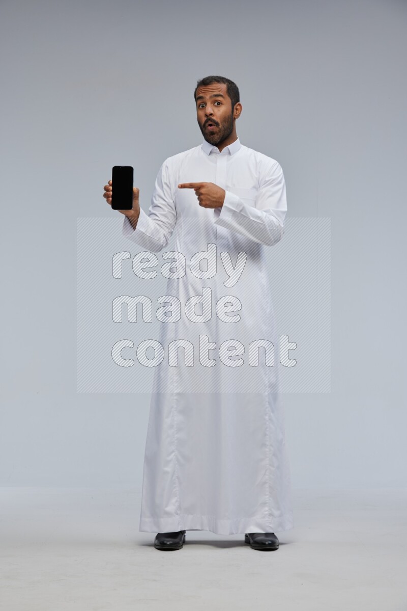 Saudi man wearing Thob standing showing phone to camera on Gray background