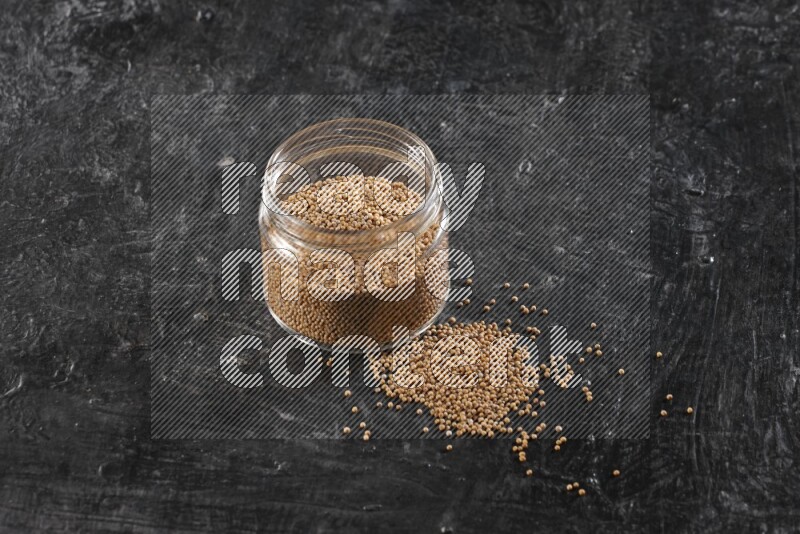 A glass jar full of mustard seeds and jar is flipped and seeds spread out on a textured black flooring