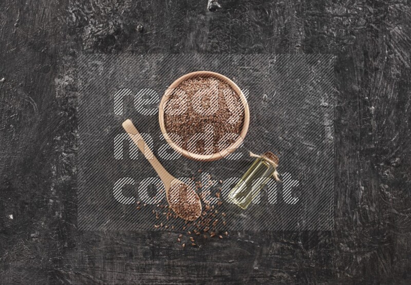 A wooden bowl and spoon full of flaxseeds and a glass bottle of flaxseeds oil on a textured black flooring