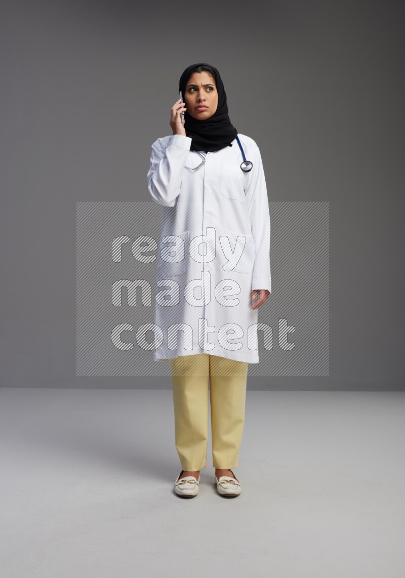 Saudi woman wearing lab coat with stethoscope standing talking on phone on Gray background