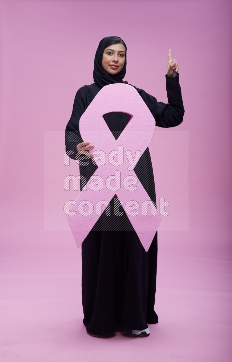 Saudi woman wearing pink ribbon on abaya standing holding awareness ribbon on plain pink background