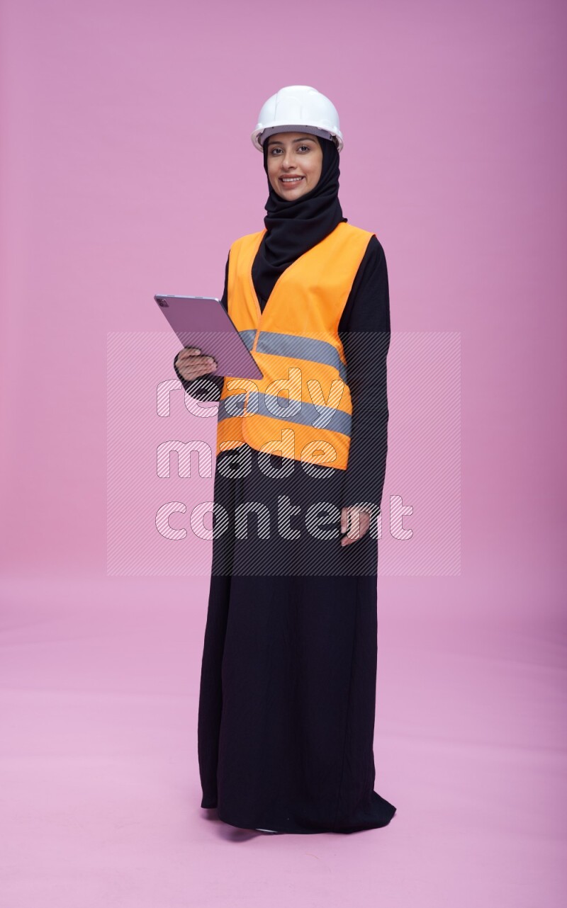 Saudi woman wearing Abaya with engineer vest and helmet standing working on tablet on pink background