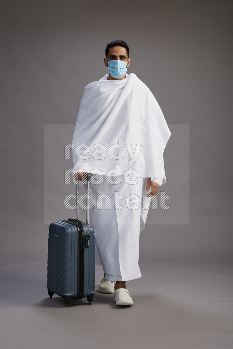 A man wearing Ehram with face mask Standing holding traveling bag on gray background