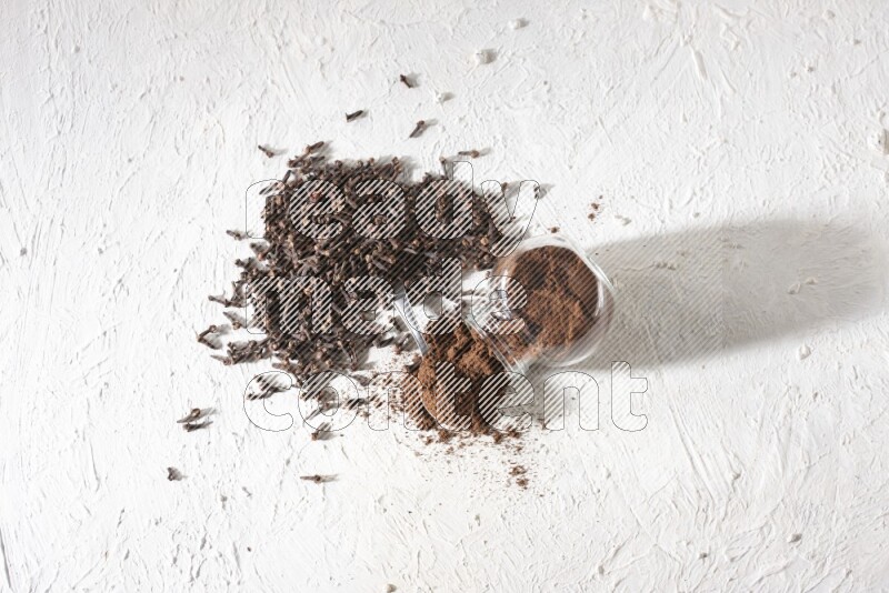 A flipped glass spice jar and a metal spoon full of cloves powder and powder came out of the jar with cloves spread on textured white flooring
