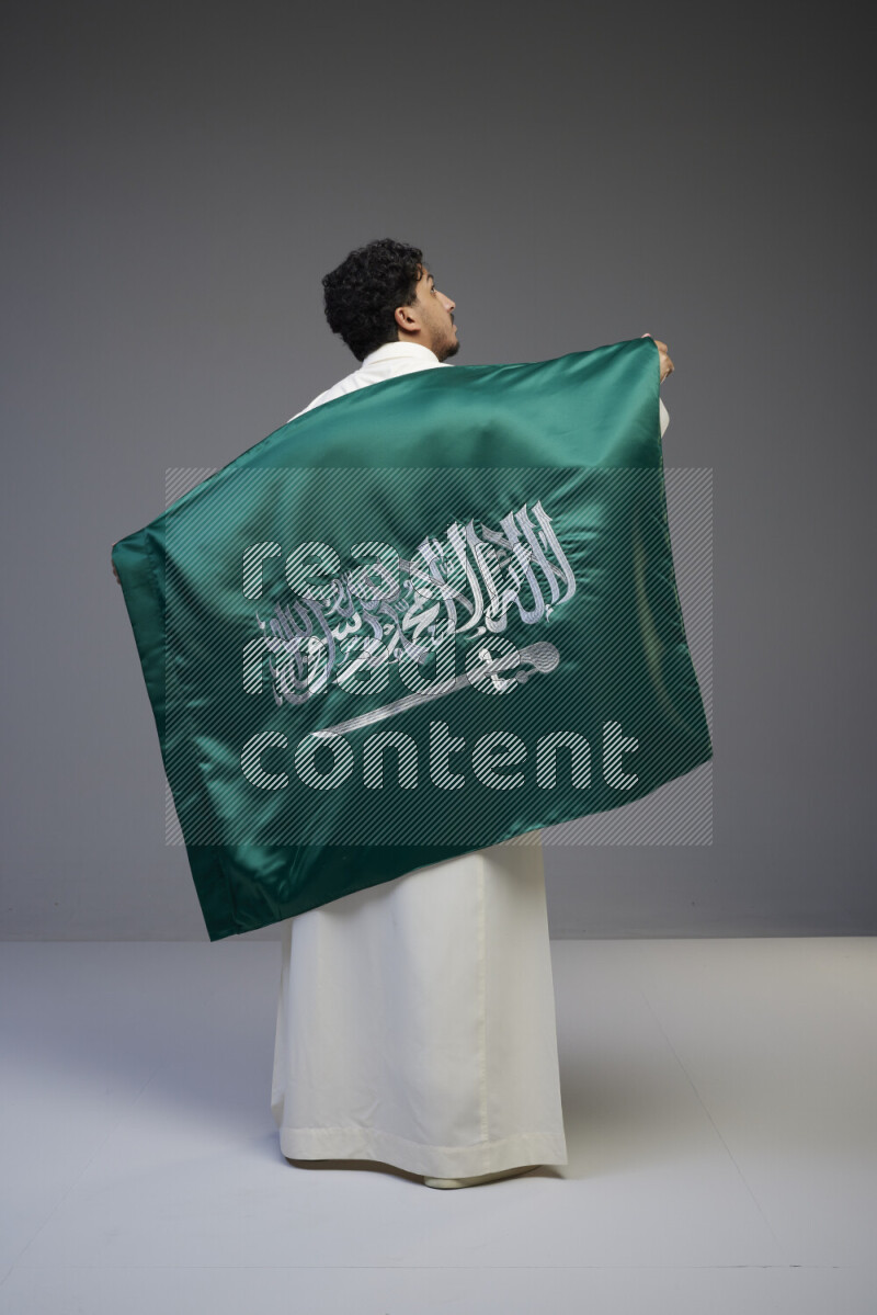 A Saudi man standing wearing thob holding big Saudi flag on gray background