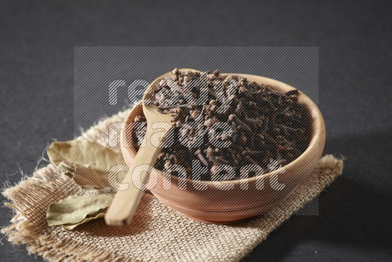 A wooden bowl, a wooden spoon full of cloves, and bay leaves (laurel) on a piece of burlap on a black flooring