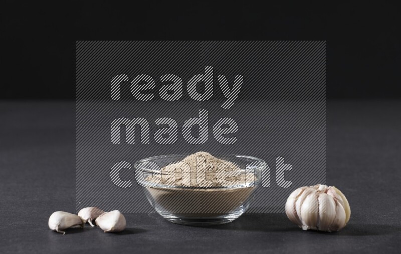 وعاء زجاجي ممتلئ ببودرة الثوم مع بعض فصوص الثوم ورأس ثوم كاملة على خلفية سوداء