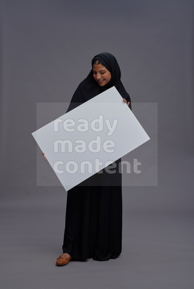 Saudi woman wearing Abaya standing holding white board on gray background