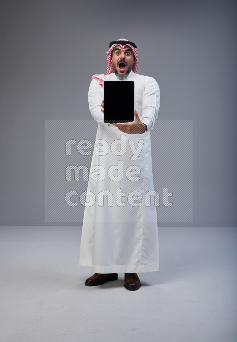 Saudi man Wearing Thob and red Shomag standing showing tablet to camera on Gray background