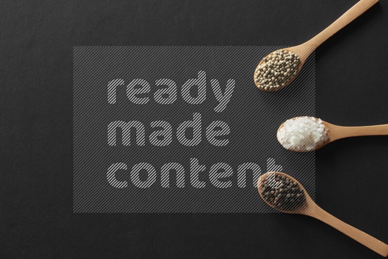 wooden spoons filled with white peppers, cloves, cardamom, salt, black peppers and basil on black flooring