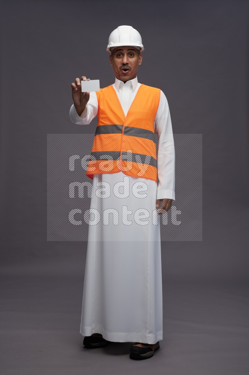 Saudi man wearing thob with engineer vest standing holding ATM card on gray background