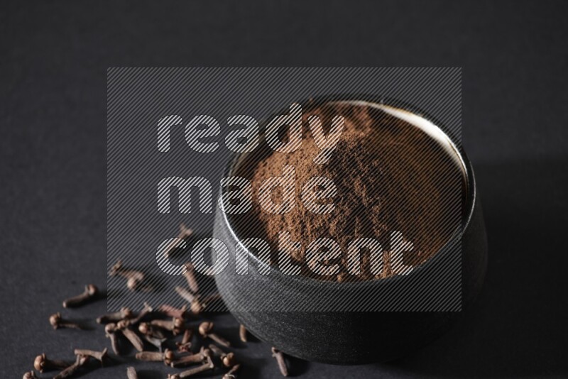 A black pottery bowl full of cloves powder and cloves whole grains on a black flooring