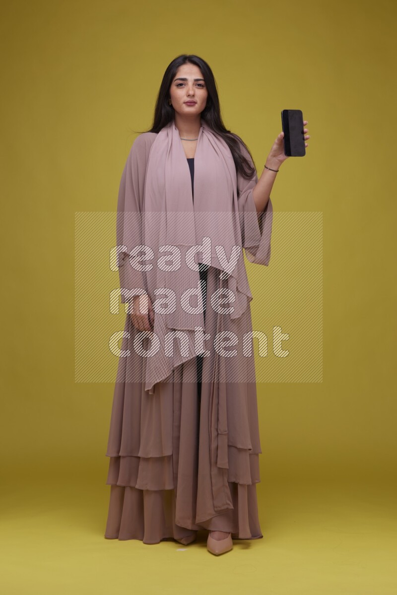 A woman Showing SmartPhone Screen on a Yellow Background wearing Brown Abaya