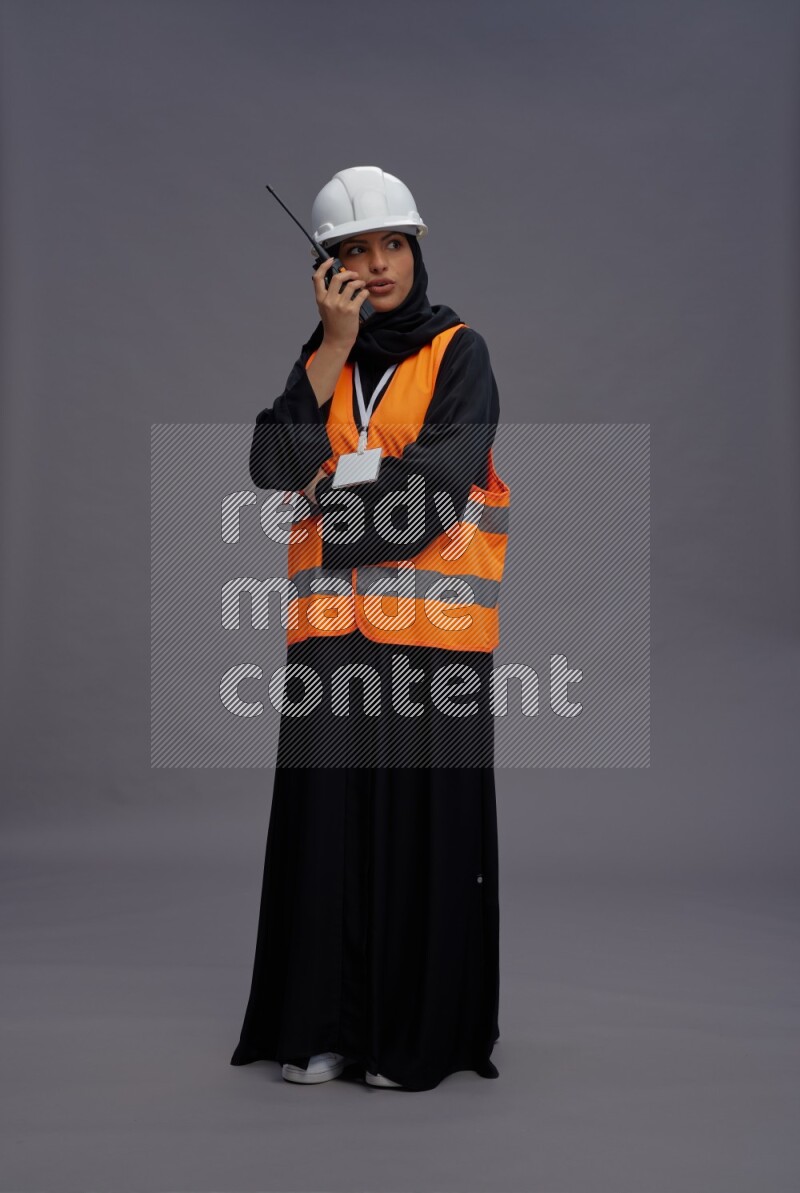 Saudi woman wearing Abaya with engineer vest with neck strap employee badge standing holding walkie-talkie on gray background
