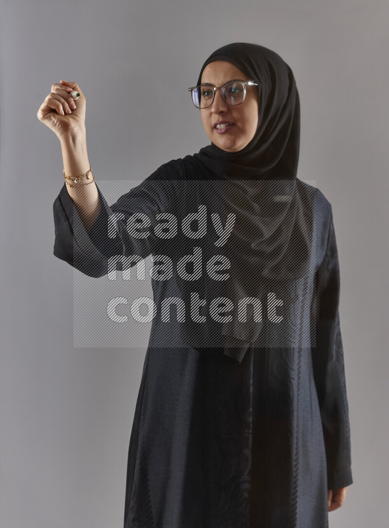 Woman in black abaya, hijab and glasses holding a marker pen to write on a transparent board with different reactions and expressions on grey background