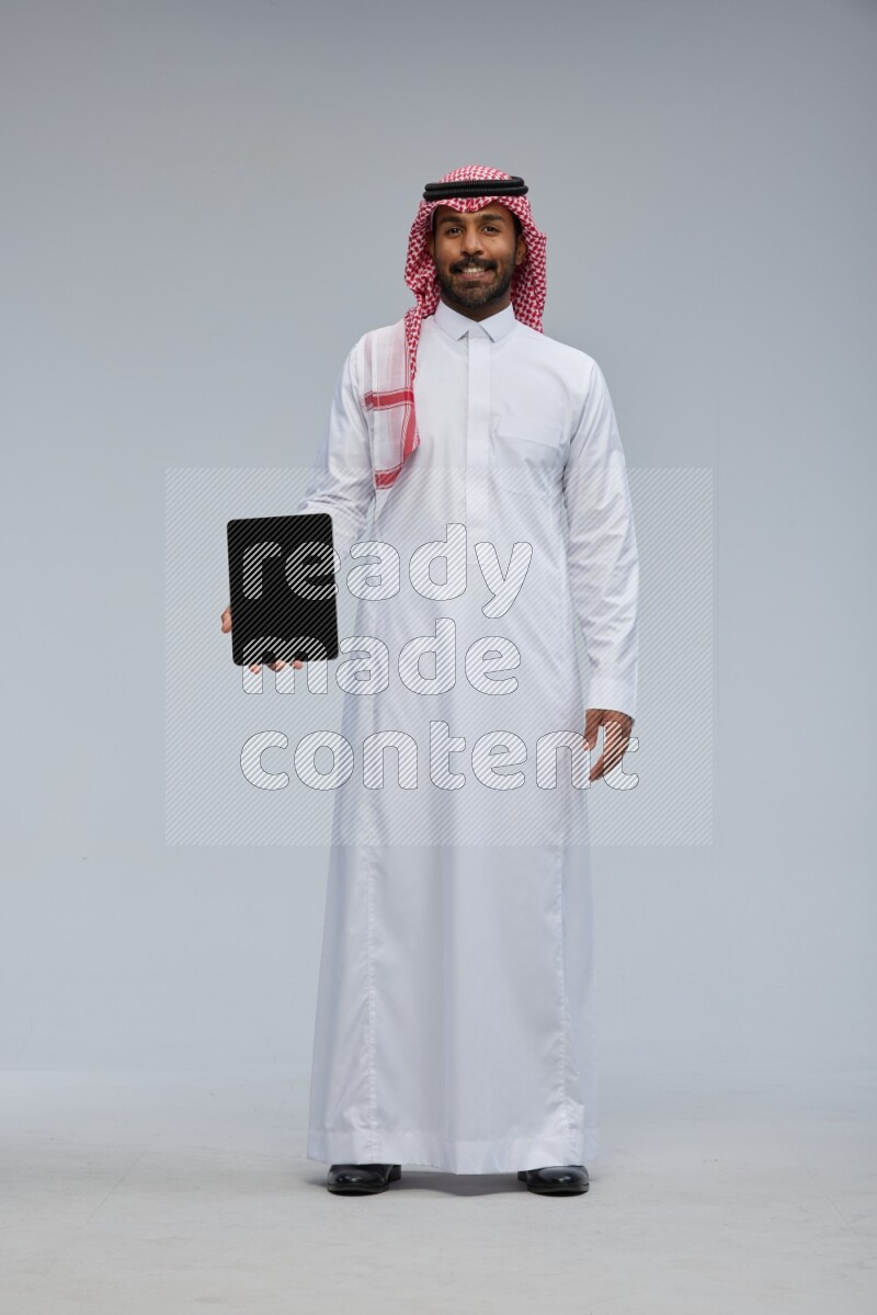 Saudi man Wearing Thob and shomag standing showing tablet to camera on Gray background