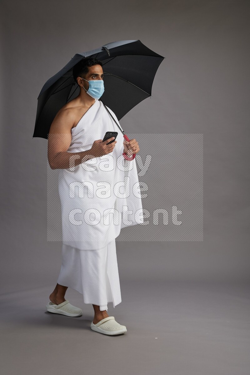 A man wearing Ehram and mask Standing holding umbrella on gray background on gray background