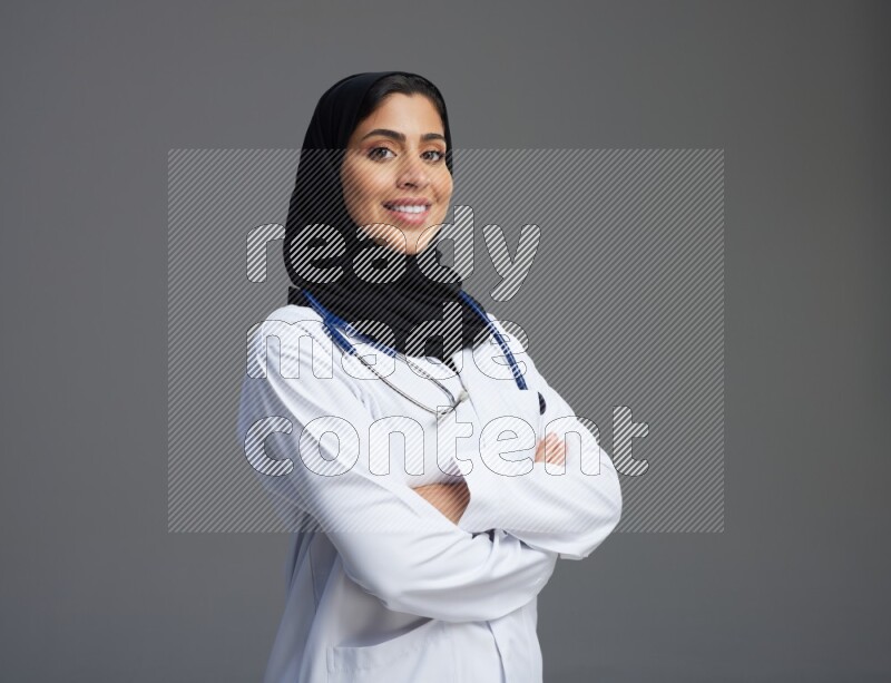 Saudi woman wearing lab coat with stethoscope standing with crossed arms on Gray background