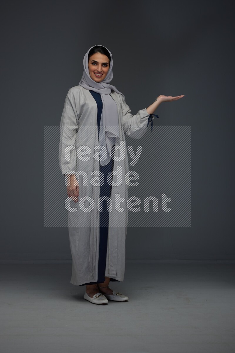A Saudi woman wearing a light gray Abaya and head scarf standing and pointing in different directions eye level on a grey background