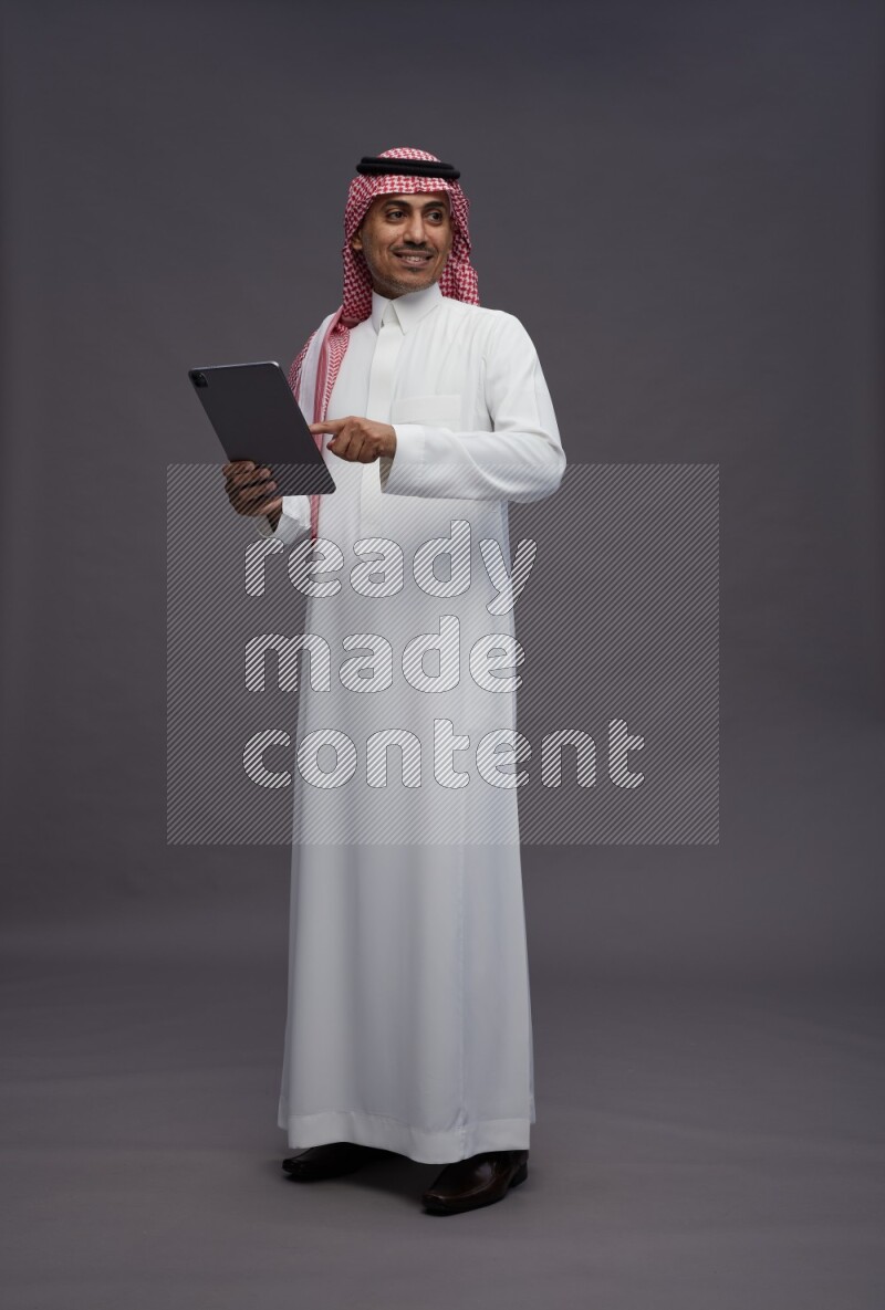 Saudi man wearing thob and shomag standing working on tablet on gray background
