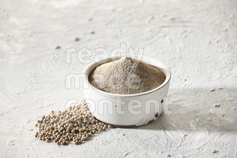 white pottery bowl full of white pepper powder with pepper beads on textured white flooring