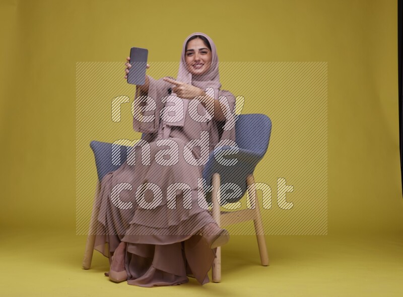 A woman Showing Her Phone Screen Setting  on a Yellow Background wearing Brown Abaya with Hijab