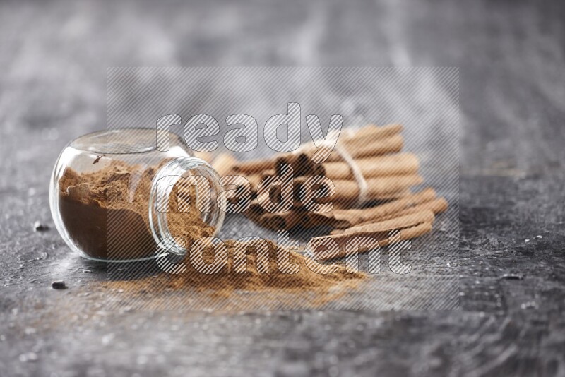 Herbal glass jar full of cinnamon powder flipped with cinnamon sticks stacked and bounded on a textured black background