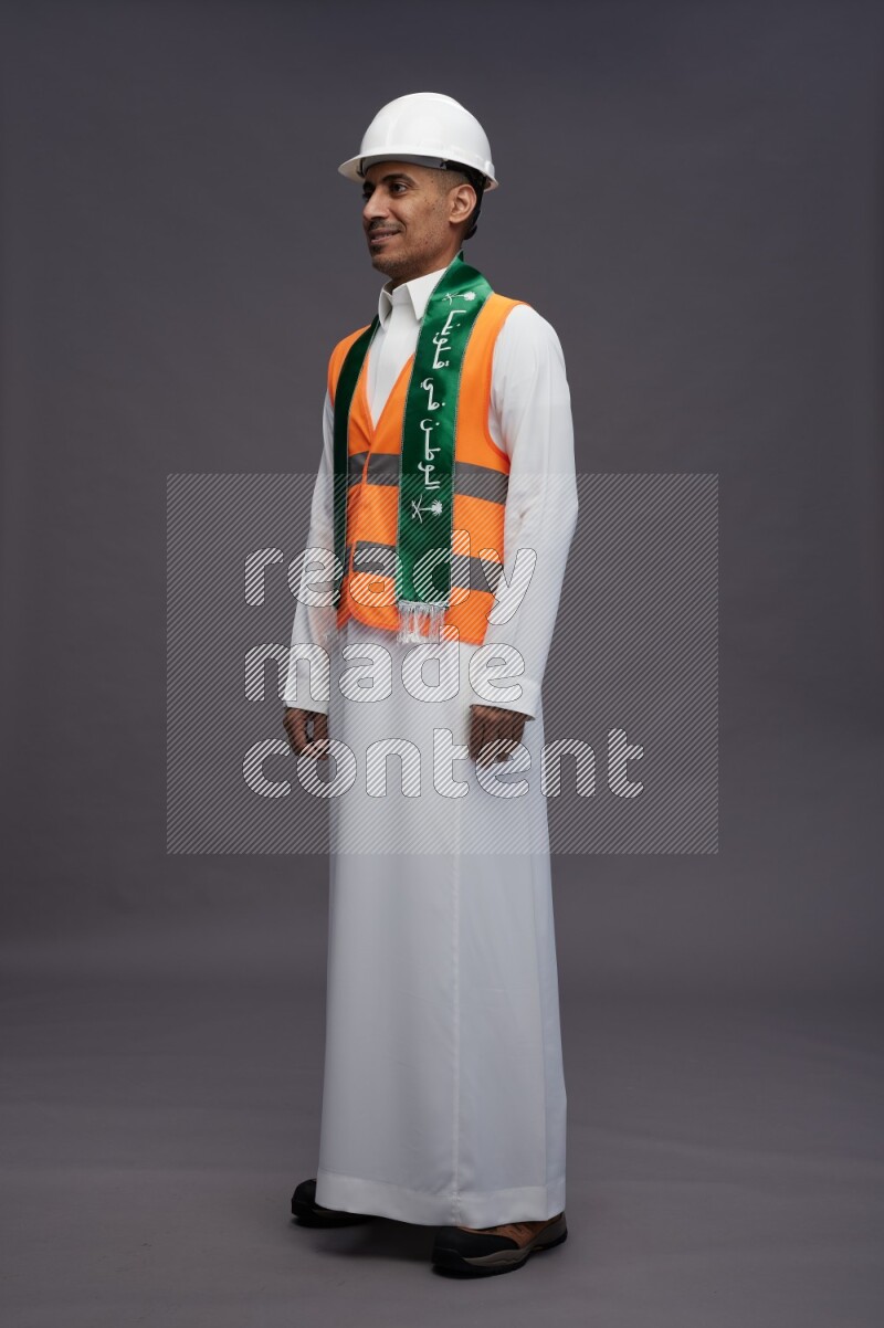 Saudi man wearing thob with engineer vest standing holding Saudi flag on gray background